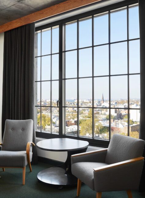 Two gray armchairs and a round table are positioned by a large window with black curtains, overlooking a cityscape.