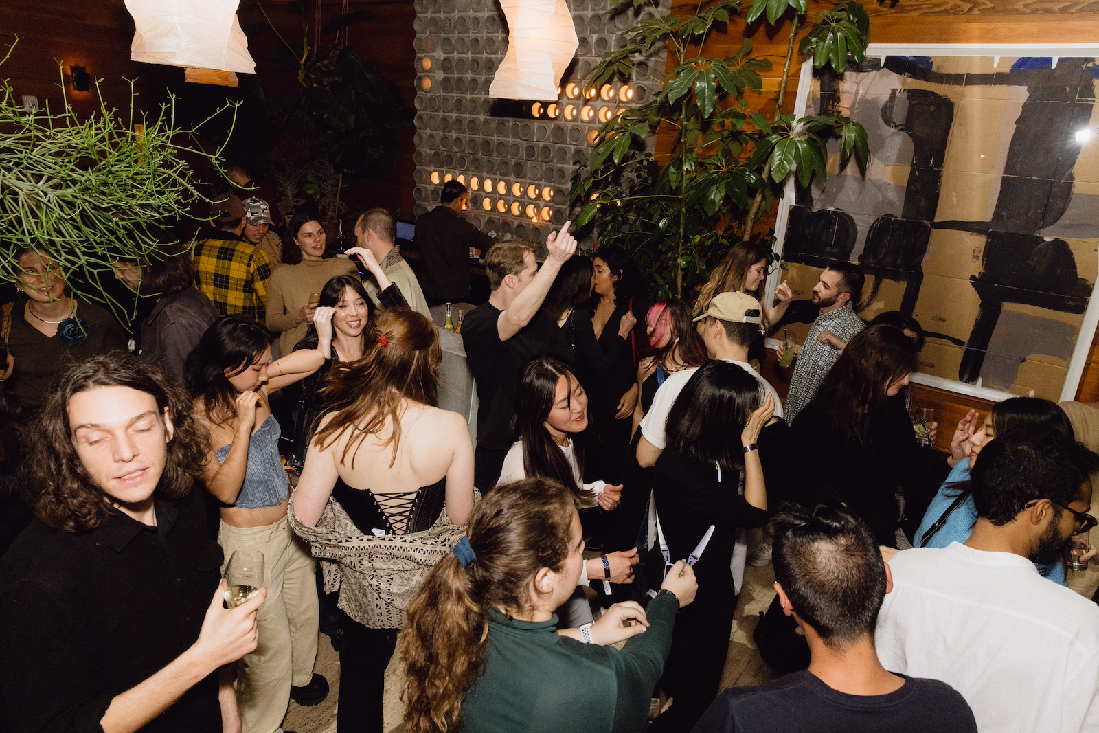 A group of people socialize and dance at an indoor party with dim lighting and plants around the room.