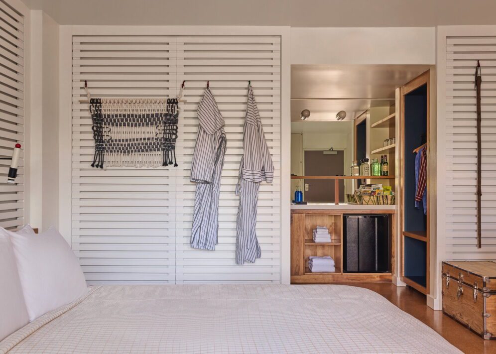 two robes hang against a slotted wall, and a large mirror sits above towels and a mini fridge