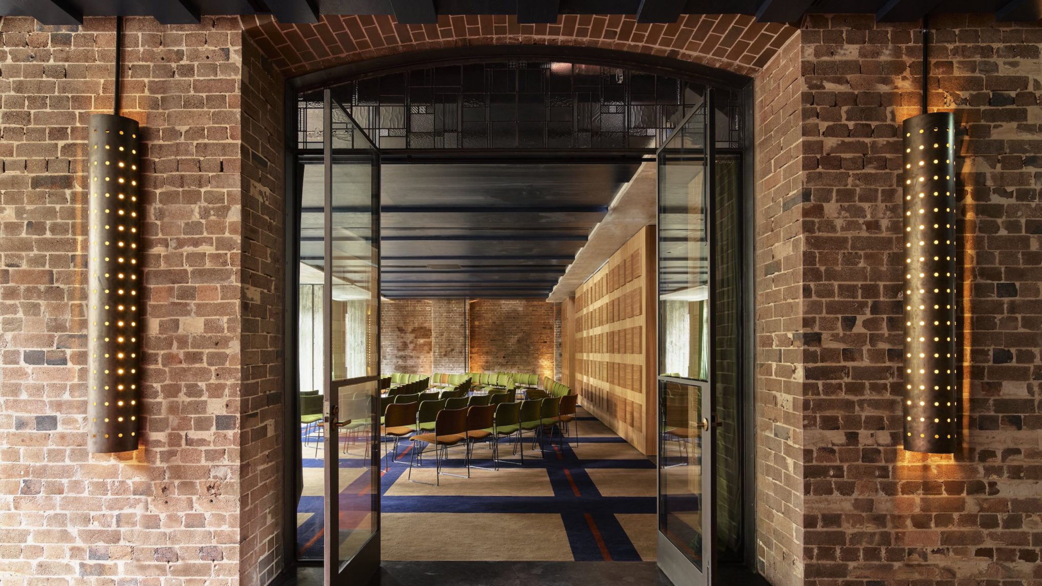 Glass doors open to a modern conference room with rows of green chairs, exposed brick walls, and patterned carpet.