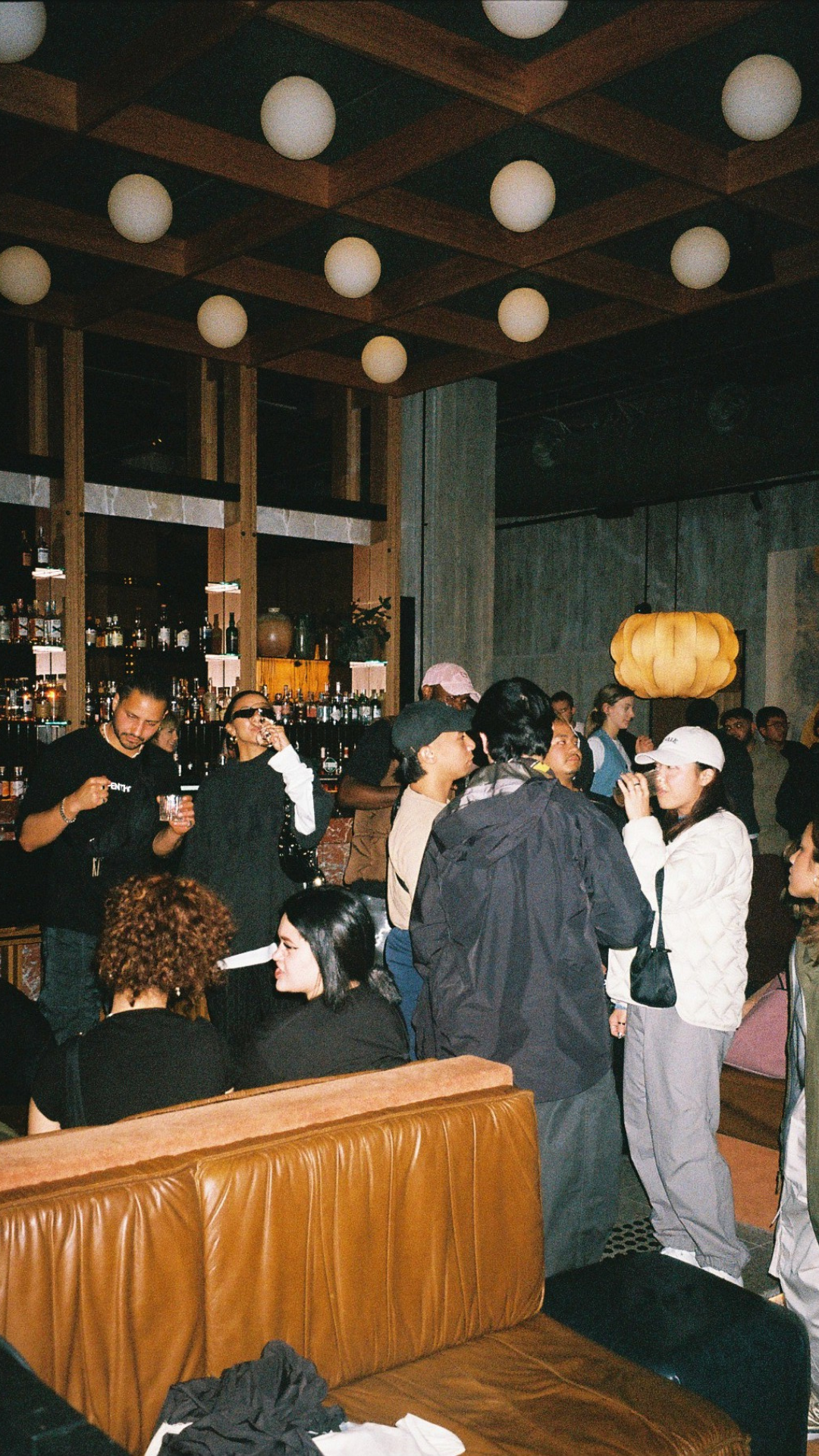 A group of people socializing and drinking in a dimly lit bar with shelves of bottles visible in the background.